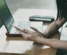 female-arms-leaning-on-table-holding-smartphone-with-open-laptop-on-table